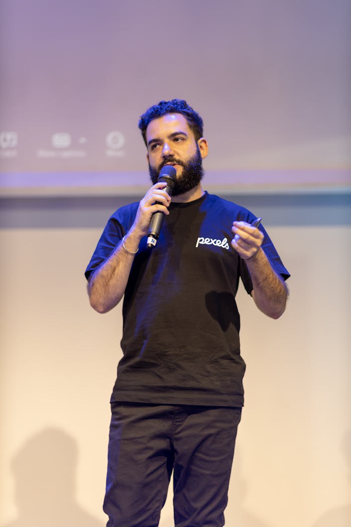 A confident speaker presenting with a microphone on stage under professional lighting.